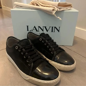 Svarta sneakers från Lanvin med lackad tå - Snygga svarta sneakers från Lanvin med klassisk siluett, snörning och vit sula. Ovandelen är i mocka och tån är i blankt lackmaterial som ger en cool kontrast. Perfekta för dig som gillar stilrena men ändå unika skor.