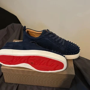 Mörkblå Christian Louboutin sneakers - Snygga mörkblå sneakers från Christian Louboutin i mocka med ikoniska röda sulor och coola nitar framtill. Skorna har snörning och vit platt sula, perfekt för dig som vill sticka ut med en lyxig och edgy stil.
