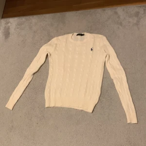 Vit kabelstickad tröja Polo Ralph Lauren - Kabelstickad vit tröja från Polo Ralph Lauren med klassisk rund hals och broderad logga på bröstet. Långärmad och i slim passform, perfekt för dig som gillar en clean och tidlös stil. Tillverkad i mjuk bomull för extra komfort. Skriv vid frågor samt intresse!