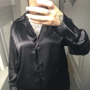 Svart silkeslen skjorta med knappar - Säljer en svart skjorta i glansigt, silkesliknande material med klassisk krage och knäppning framtill. Skjortan har lång ärm och en loose passform som ger en avslappnad men ändå stilren look. Perfekt för dig som gillar en clean och modern vibe. Passar S-M