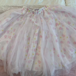 Söt kortkjol med kaninmotiv från Kawaii.se - Supergullig kortkjol från Kawaii.se med vitt tyg, rosa kaniner och hjärtan i rosa och gult. Kjolen har ett lager av tunn tyll ovanpå och resår i midjan för extra komfort. Perfekt för dig som älskar kawaii-stil och vill sticka ut med ett lekfullt plagg. Den är i storlek S och passar perfekt som en söt underkjol!!<3