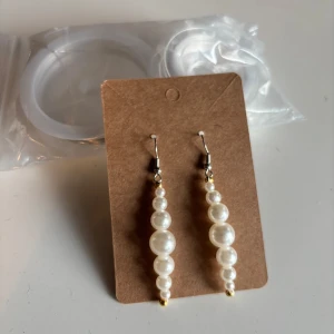 Långa pärlörhängen med vita pärlor - Snygga örhängen med hängande vita pärlor i olika storlekar på rad. Krokar i silverfärg och små gulddetaljer längst ner. Perfekta för dig som vill ha en elegant och trendig look med klassiska pärlor.