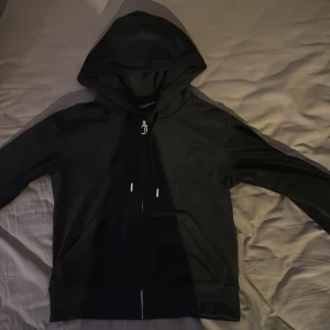 Svart hoodie från Juicy Couture - Svart hoodie från Juicy Couture med dragkedja och huva. Klassisk design med fickor framtill och snörning i huvan. Liten broderad logga på bröstet och mjukt material.