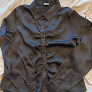 Svart satinblus med knappar - Snygg svart blus i satin med klassisk krage och knappar framtill. Långärmad modell med lätt glansigt material och rynkade detaljer längs knappraden. Perfekt för dig som gillar stilrena och trendiga plagg.