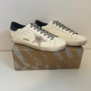 Golden Goose vita sneakers med stjärna - Snygga vita sneakers från Golden Goose med grå stjärndetalj på sidan, grå snörning och svart häl. Skorna har en vintage-inspirerad look med slitna detaljer och platt sula. Perfekta för dig som gillar streetstyle och unika detaljer.