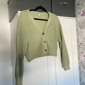 Ljusgrön stickad kofta från Monki - Superfin ljusgrön stickad kofta från Monki i croppad modell. Koftan har v-ringning, stora knappar framtill och långa ärmar. Passar XS-L