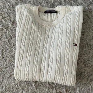 Vit kabelstickad tröja Tommy Hilfiger - Vit kabelstickad tröja från Tommy Hilfiger med rund halsringning och diskret logga på bröstet. Tröjan har lång ärm och ribbade muddar, perfekt för en clean och klassisk look. Materialet är mjukt och känns lyxigt mot huden.