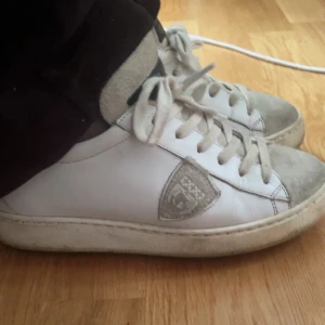 Vita Philippe Model sneakers i skinn - Snygga vita sneakers från Philippe Model Paris med beige detaljer och klassisk sköldlogga på sidan. Skorna har snörning, rund tå och är tillverkade i skinn med mockadetaljer på tån och plösen. Perfekta för en clean och stilren look. Pris går diskuteras vid snabb affär. Går defensivt putsa upp.