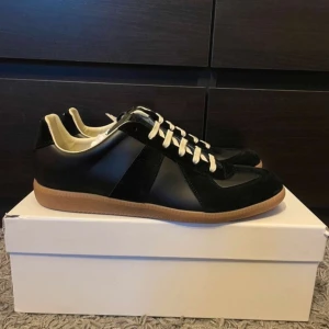 Svarta Maison Margiela Gats - Maison Margiela sneakers i svart skinn och mocka med beige innersida och vita snören. Klassisk siluett med gummisula i naturfärg och diskret branding på plösen. Perfekta för dig som gillar stilrena och tidlösa sneakers med premiumkänsla. Skorna är 10/10 skick och allt OG följer med dvs kvitto, box och dustbags. Vid frågor eller funderingar hör av dig👍