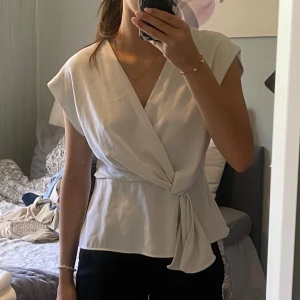 Vit omlottopp från Zara - Snygg vit omlottopp från Zara med knyt i midjan och v-ringning. Toppen är ärmlös och har en lätt glansig yta som ger en lyxig känsla. Den har en liten smink fläck som borde gå bort med fläckborttagning där av priset.