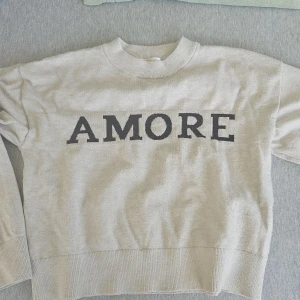 Beige sweatshirt AMORE Gina Tricot - Beige sweatshirt från Gina Tricot med svart texten 'AMORE' framtill. Tröjan har rund halsringning, ribbade muddar och är långärmad. Perfekt för en chill och trendig look.