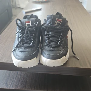Svarta Fila Disruptor sneakers - Chunky svarta Fila Disruptor sneakers med vit, kraftig sula och snörning. Skorna har Fila-logga på sidan, plösen och hälen. Tillverkade i syntetmaterial med rund tå och platt sula. Perfekta för dig som gillar streetstyle och vill sticka ut.