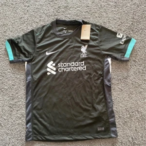 Liverpool FC svart Nike fotbollströja - Snygg svart Liverpool FC fotbollströja från Nike med vita loggor och sponsortryck på bröstet. Tröjan har korta ärmar med turkosa detaljer och grå sidopaneler. Tillverkad i lätt och ventilerande material, perfekt för träning eller match.