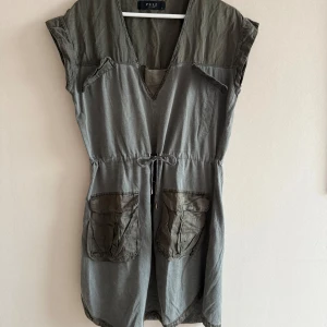 Grå/ grön klänning med snörning från Pulz jeans  - Snygg grå/ grön kortklänning från Pulz jeans  med v-ringad hals och coola detaljer som snörning i midjan och två stora fickor framtill. Klänningen är ärmlös och har en loose passform, perfekt för dig som gillar avslappnad stil. Materialet är mjukt. Rökfritt hem.  