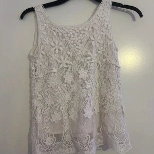 Vit spetslinne med blommigt mönster - Superfint vitt linne med blommig spets och genombrutet mönster. Ärmlös modell med v-ringning och romantiska detaljer. Perfekt att styla med jeans eller kjol för en fräsch look.