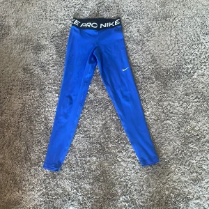 Blå Nike Pro leggings - Snygga blå Nike Pro leggings med svart midjeresår där det står 'NIKE PRO' i vitt. Tight passform och klassisk Nike-logga på benet. Perfekta för träning eller sportiga outfits.