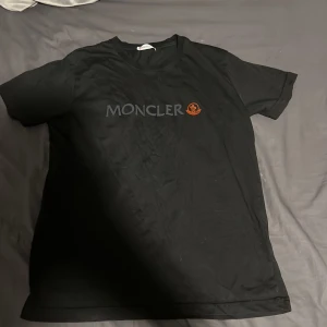 Svart Moncler t-shirt med logga - Svart t-shirt från Moncler i klassisk modell med rund hals och korta ärmar. Framsidan pryds av Moncler-loggan i svart och en röd detalj. T-shirten är gjord i mjuk bomull och har en clean, stilren look som passar perfekt till jeans eller shorts.