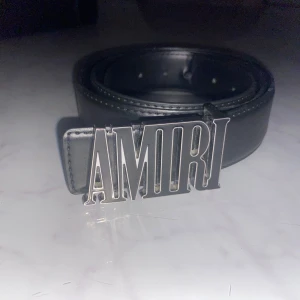 Svart bälte från Amiri  - Säljer ett svart bälte från Amiri med en stor, stilren silverspänne med AMIRI-loggan. Bältet har klassisk design och kommer med originalförpackning, dustbag och certifikat. Perfekt accessoar för att lyfta din outfit.