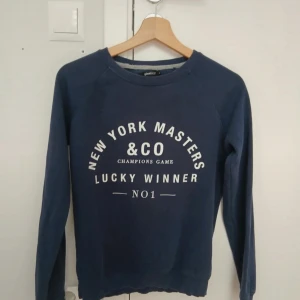 Mörkblå sweatshirt från Gina Tricot - Mörkblå sweatshirt från Gina Tricot med vit text på bröstet: 'New York Masters &Co Champions Game Lucky Winner No1'. Klassisk rund halsringning och långa ärmar. Perfekt för en chill och sportig look.