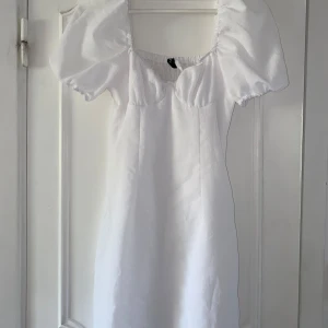 Vit kort klänning med puffärmar - Supersöt vit klänning från H&M med puffiga korta ärmar och fyrkantig ringning. Smockad rygg för extra komfort och snygg passform. Perfekt för dig som gillar en feminin och trendig stil. Materialet känns lätt och luftigt, perfekt för varma dagar.