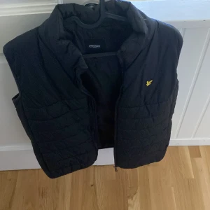 Svart dunväst från Lyle & Scott Junior - Svart quiltad dunväst från Lyle & Scott Junior med gul logga på bröstet. Västen har hög krage och dragkedja framtill. Perfekt för lager-på-lager och har en clean, sportig look som funkar till många stilar.