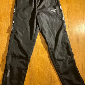 Svarta träningsbyxor från Umbro - Svarta träningsbyxor från Umbro med vita loggor längs sidorna och broderad Umbro-logga fram. Byxorna har resår i midjan och är gjorda i ett lätt, glansigt polyestermaterial som andas. Perfekta för sport eller chill.