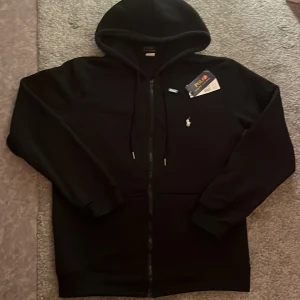 Svart hoodie från Polo Ralph Lauren - Svart hoodie från Polo Ralph Lauren med klassisk huva och dragkedja framtill. Tröjan har den ikoniska Polo-loggan broderad i vitt på bröstet och är tillverkad i 100% polyester. Långärmad modell med ribbade muddar och snörning vid huvan. Perfekt för chill dagar.