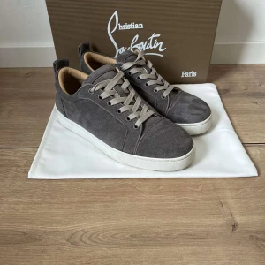 Grå mocka sneakers Christian Louboutin - Snygga grå sneakers från Christian Louboutin i mjuk mocka med vita platta sulor och ljusgrå snörning. Skorna har en rund tå och beige innersida som ger en lyxig känsla. Perfekta för dig som gillar stilrena och exklusiva sneakers.