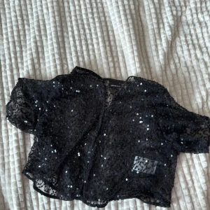 Svart paljett bolerokofta från Gina Tricot - Säljer en svart transparent bolerokofta från Gina Tricot, täckt med glittrande paljetter. Toppen har korta ärmar, croppad passform och är gjord i ett lätt syntetmaterial med mönstrad mesh. Perfekt att slänga över ett linne för en extra touch.