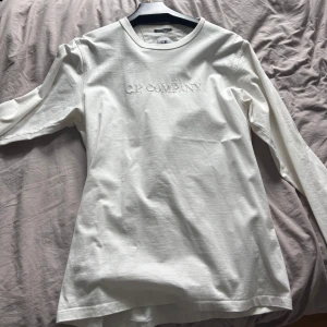 Vit långärmad tröja från C.P. Company - Säljer en stilren vit långärmad tröja från C.P. Company med broderad logga på bröstet. Tröjan har en klassisk rund hals och är gjord i mjuk bomull som känns skön mot huden. Perfekt för dig som gillar minimalistisk design och snygga detaljer.