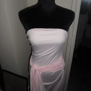 Ljusrosa axelbandslös långklänning - Säljer en elegant ljusrosa axelbandslös långklänning med mjukt fall och knyt i midjan. Klänningen har en slät yta och är gjord i ett glansigt, följsamt material som ger en snygg siluett. 