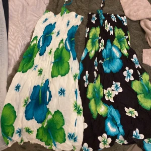 Blommiga sommarklänningar med smock - Två korta klänningar med tropiskt blommönster i blått, grönt och vitt. Ena klänningen har vit botten, den andra svart. Båda har smock upptill och knyt runt halsen. Perfekta för varma dagar och har en luftig, avslappnad passform. Står inget märke. 40kr/st