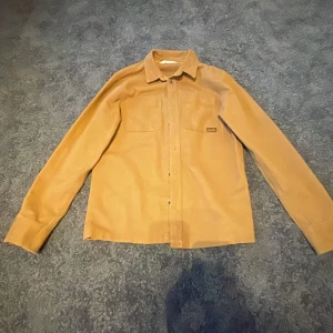 Beige overshirt från H&M NYC/93 - Snygg beige overshirt från H&M med klassisk krage, två bröstfickor och cool NYC/93-detalj på ena fickan. Skjortan har långa ärmar och knäppning framtill. Perfekt att slänga över en t-shirt för en chill look.