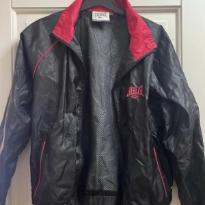 Svart och röd vindjacka från Everlast - Snygg svart vindjacka från Everlast med röda detaljer och logga på bröstet. Jackan har meshfoder, dragkedja och röda kontrastsömmar. Väldigt bra skick, inga defekter! 🤗Står som storlek L men passar allt från L till Xs. Jag brukar ha xs och S och den passar mig fint🥰