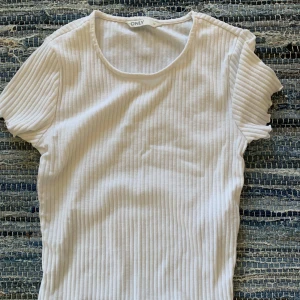 Vit ribbad topp från ONLY - En stilren vit ribbad topp från ONLY med rund halsringning och korta ärmar. Toppen har en smickrande passform och är tillverkad i mjukt bomullsmaterial. Perfekt att matcha med jeans eller kjol för en clean och fräsch look.