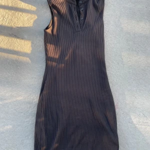 Svart ribbad kortklänning Divided - Snygg svart ribbad kortklänning från Divided med ståkrage och knappdetaljer framtill. Ärmlös design och figurnära passform som ger en cool och modern look. Perfekt för dig som gillar minimalistisk stil och vill ha något enkelt men trendigt. Den är i storlek XXS men jag brukar använda Xs och S så skulle säga det mer sitter som en Xs