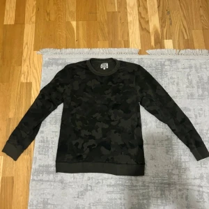 Crocker camo sweatshirt i mörkgrön - Snygg mörkgrön sweatshirt från Crocker med camouflagemönster. Tröjan har rund halsringning och långa ärmar, perfekt för dig som gillar streetstyle. Tillverkad i mjuk bomullsmix som är skön att ha på sig hela dagen.