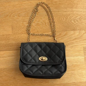 Svart quiltad axelväska med guldkedja - Snygg svart axelväska med quiltat mönster och guldfärgad kedja som axelrem. Väskan har ett klassiskt lock med vridspänne i guld och är tillverkad i syntetmaterial. Perfekt storlek för det viktigaste och ger en chic vibe till din outfit.