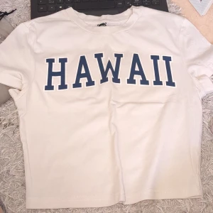 Vit t-shirt med HAWAII-tryck - Snygg vit t-shirt med stort blått HAWAII-tryck på bröstet. Klassisk rund hals och korta ärmar, perfekt för chill dagar. T-shirten har en rak passform och är gjord i mjuk bomull som känns skön mot huden.