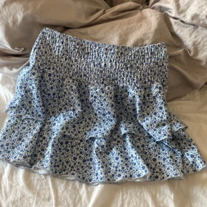 Blommig volangkjol från Gina Tricot - Supersöt kort kjol från Gina Tricot med blått blommönster på vit botten. Kjolen har smock i midjan och flera volanger som ger en flowy och lekfull look. Perfekt för dig som gillar romantisk stil och vill ha något som sticker ut lite extra.