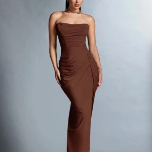 Brun axelbandslös långklänning - Snygg brun axelbandslös långklänning med draperade detaljer och slits på sidan. Klänningen har en figurnära passform och är tillverkad i ett mjukt, stretchigt material som sitter bekvämt mot kroppen. Perfekt för dig som vill ha en elegant och trendig look.