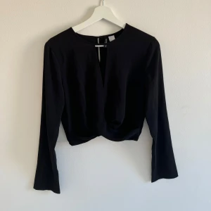 Svart croppad blus från H&M - Svart blus från H&M med croppad passform och lång ärm. Toppen har en snygg v-ringad detalj framtill med en liten rem över öppningen. Materialet är lätt strukturerat och känns mjukt mot huden. Perfekt för dig som gillar stilrena och enkla plagg.