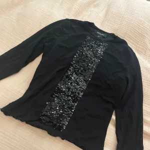 Svart paljett-tröja från Ideology - Svart långärmad tröja från Ideology i nylon med en bred, glittrig paljettpanel framtill. Tröjan har en rund hals, trekvarts armar och vågiga kanter vid nederdelen 💞. 