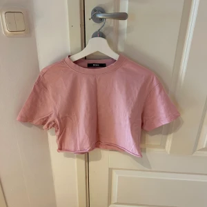 Rosa croppad topp från Bik Bok - Säljer en ljusrosa croppad topp från Bik Bok i mjuk bomull. T-shirten har rund hals, korta ärmar och loose passform. Perfekt att styla med höga jeans eller shorts för en chill look. Enkel och clean design utan mönster eller detaljer.