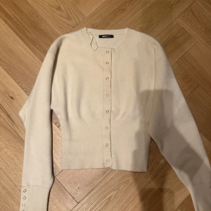 Beige kofta från Gina Tricot - Snygg beige kofta från Gina Tricot med rund hals och knappar framtill. Modellen är croppad och har långa ärmar med knappar vid ärmsluten. Perfekt att styla med jeans eller kjol för en chill look.