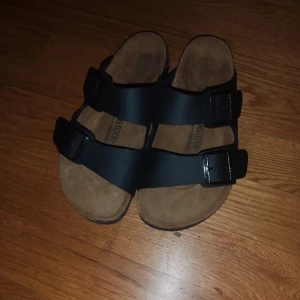 Svarta Birkenstock sandaler strl 39 - Säljer ett par svarta Birkenstock sandaler med två breda remmar och justerbara spännen. Sulan är i kork och gummi, och ovandelen är i syntetmaterial. Perfekt för dig som gillar klassisk stil och vill ha bekväma skor med platt sula.