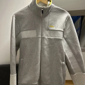 Grå ziptröja från BOSS med vita detaljer - Snygg ljusgrå ziptröja från BOSS med vita ribbade paneler på bröstet och ärmarna. Tröjan har hög krage, hel dragkedja och fickor på sidorna. BOSS-logga i gult på bröstet ger en clean och modern look. Perfekt för dig som gillar stilrena streetwear-vibbar.