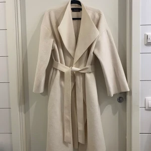 Beige kappa från Bianca x Nelly, stl 38 - Snygg beige kappa från Bianca x Nelly i storlek 38. Lång modell med bred krage och bälte i midjan för en clean och stilren look. Perfekt för dig som gillar minimalistisk stil och vill ha en jacka som sticker ut på ett classy sätt.