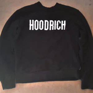 Svart Hoodrich sweatshirt med tryck - Svart sweatshirt från Hoodrich med stort vitt tryck på ryggen. Klassisk crewneck-modell med ribbade muddar vid ärmslut och midja. Perfekt för dig som gillar streetwear och vill sticka ut med en bold logga.