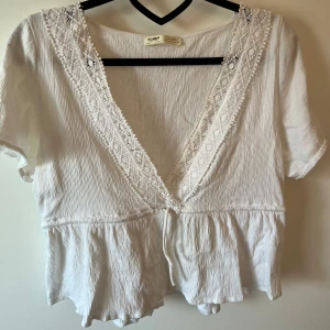 Vit blus med spets från Pull&Bear - Superfin vit blus från Pull&Bear med djup v-ringning och spetsdetaljer längs urringningen. Blusen har korta ärmar, knyt framtill och volang nedtill. Materialet är lätt och tunt. Bysten blir väldigt framhävande och snyggt.